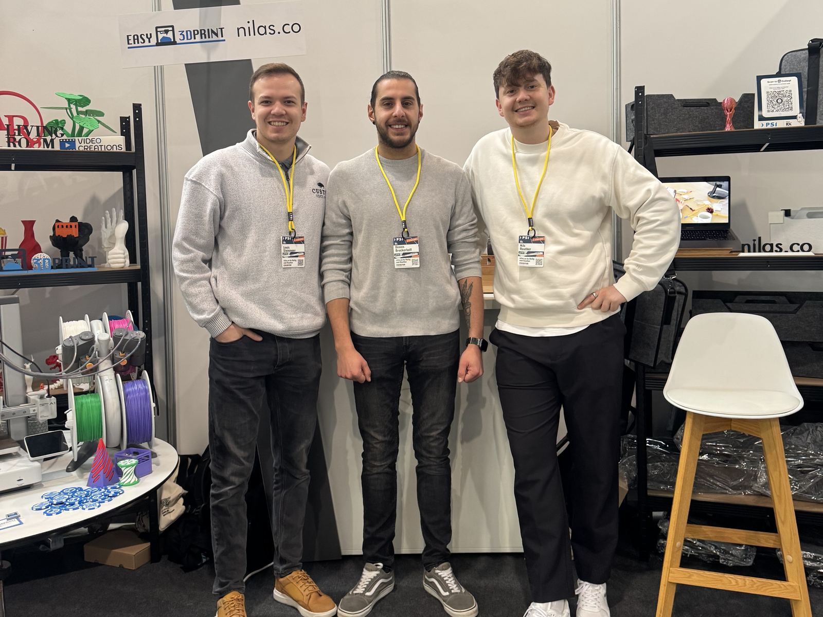 Louis, Florian und Nils von Easy 3DPrint am eigenen Messestand, daneben das Regal mit gedruckten Objekten und der Tisch mit laufender Produktion